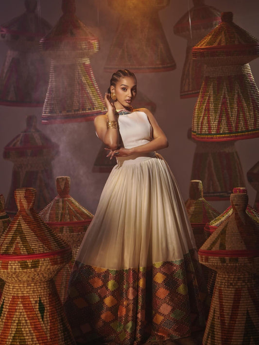 A woman models a sleeveless, high-neck Habesha Dress with a solid cream upper bodice and a voluminous skirt featuring a multicolor geometric pattern along the hem. She is posed in a stylized studio setting surrounded by hanging woven rugs.