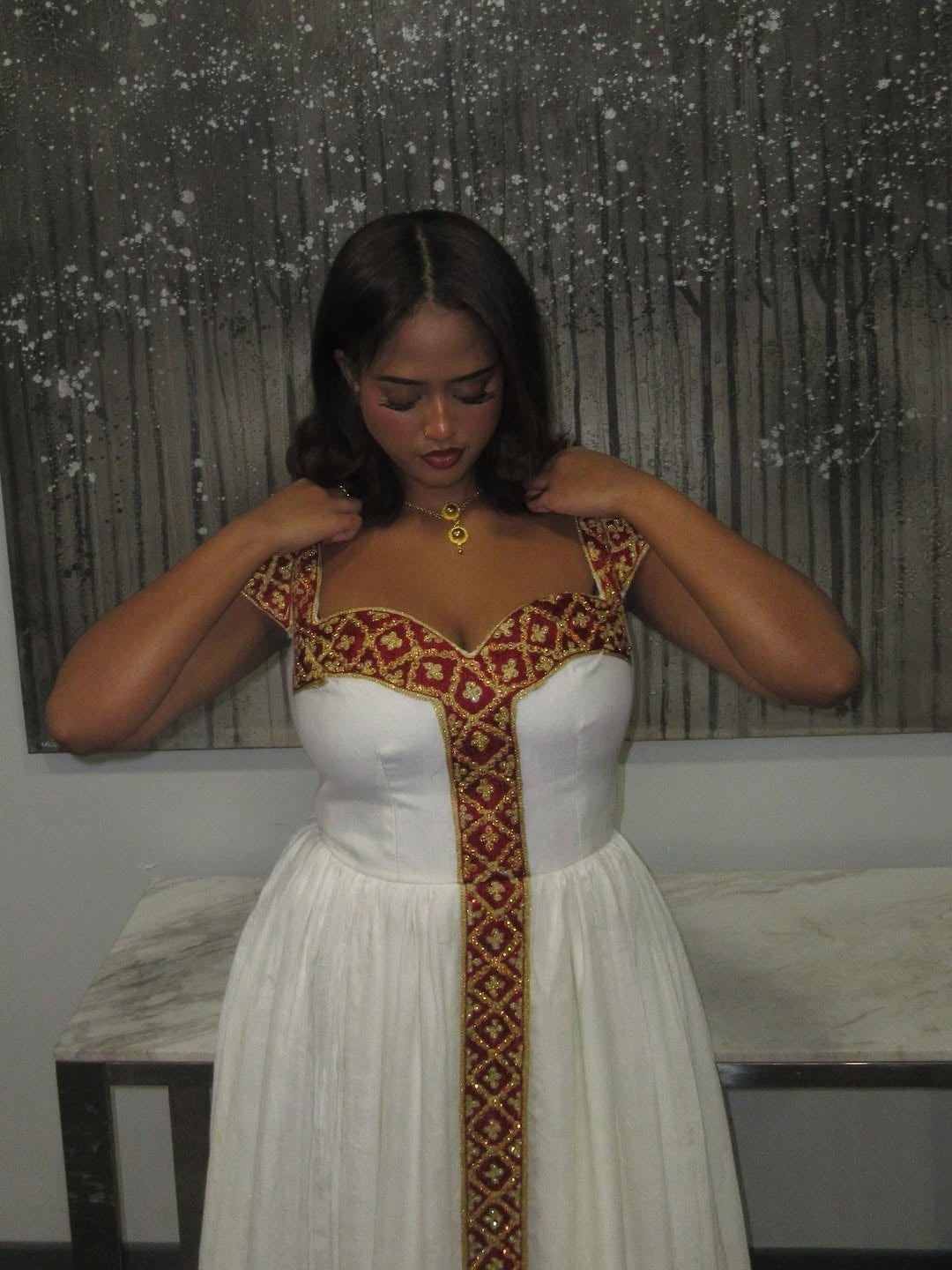 Woman wearing a white habesha kemis with red and gold embroidery in a room.