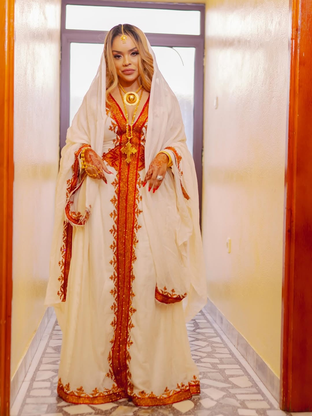 Woman in a Habesha Zuria with white and orange design standing in a hallway.