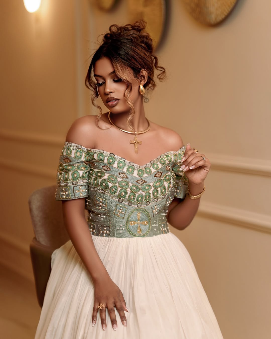 Woman wearing an off-shoulder dress with intricate green patterns in a warm-toned room.
