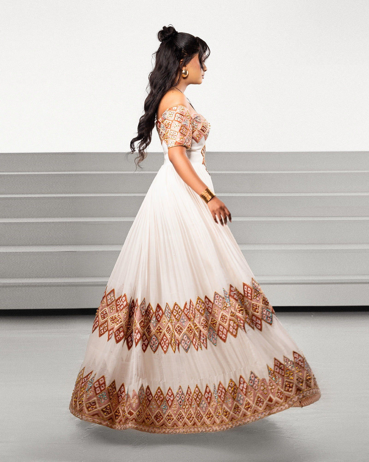 A woman modeling a floor-length off-the-shoulder Ethiopian Traditional Clothing with a cream base and extensive rust red, orange, and gold geometric embroidery concentrated on the bodice and hem, shown against a minimalist gray studio background.