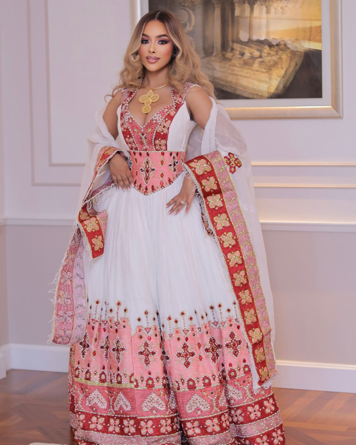 A woman models a traditional white and pink embroidered habesha Wedding dress with red and gold patterns, a matching sheer dupatta, and a gold necklace in an indoor setting.