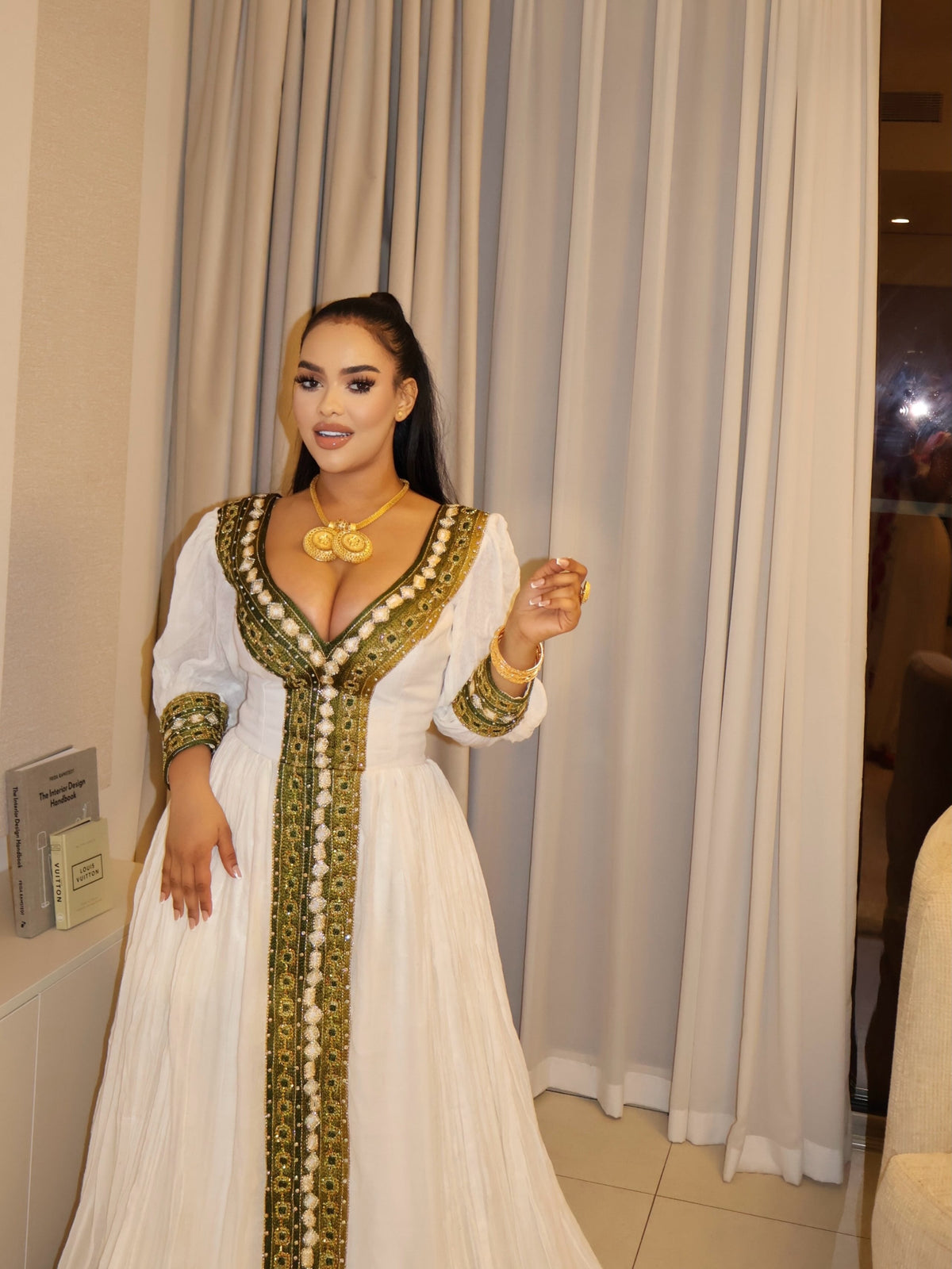 A woman modeling a white traditional long Ethiopian National Dresses with ornate gold and green embroidery, standing in front of beige curtains.