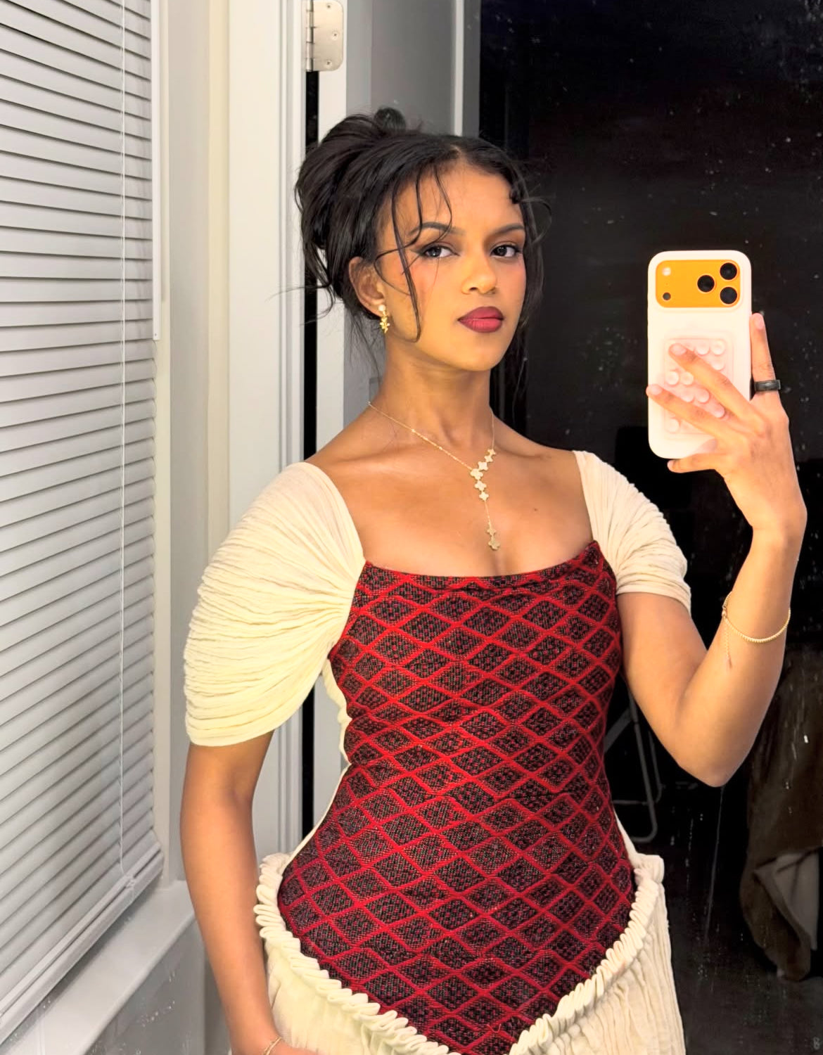 Woman taking a mirror selfie wearing a red and black Ethiopian Dress patterned top with a white cardigan.