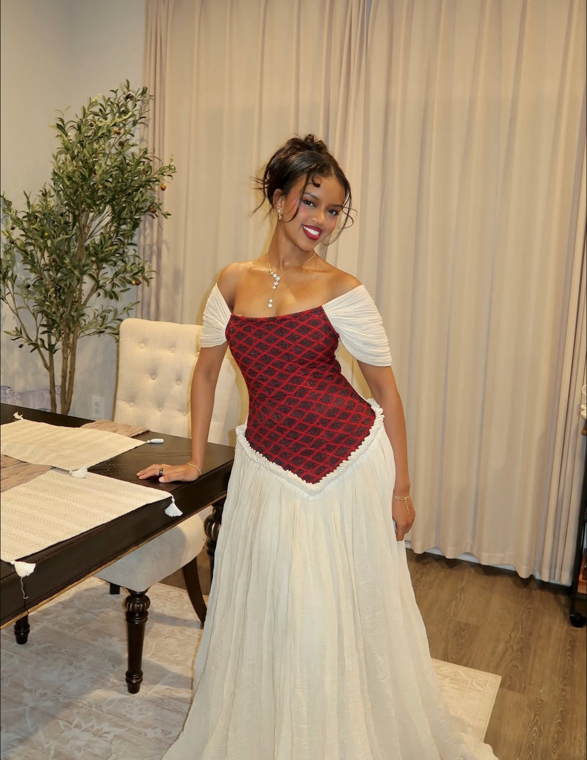 Woman in a red and white Ethiopian Dress  standing in a room with a plant and furniture.