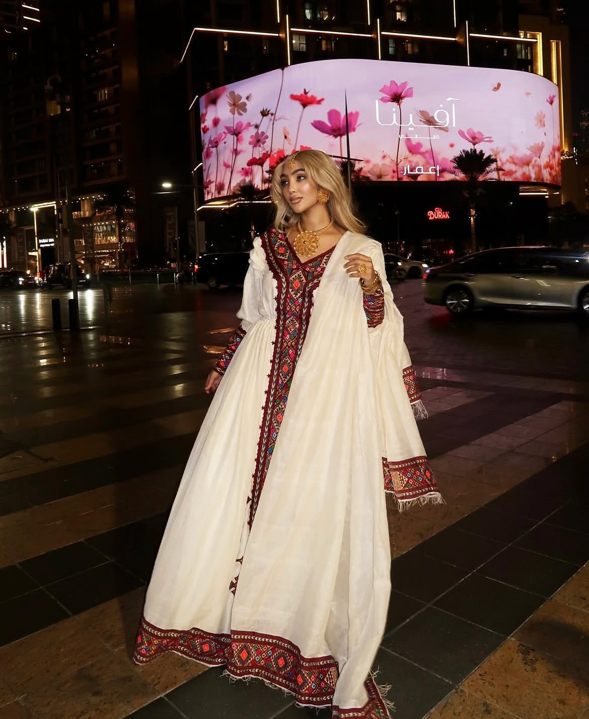 Woman in a traditional outfit standing on a city street at night with a large screen displaying flowers in the background.