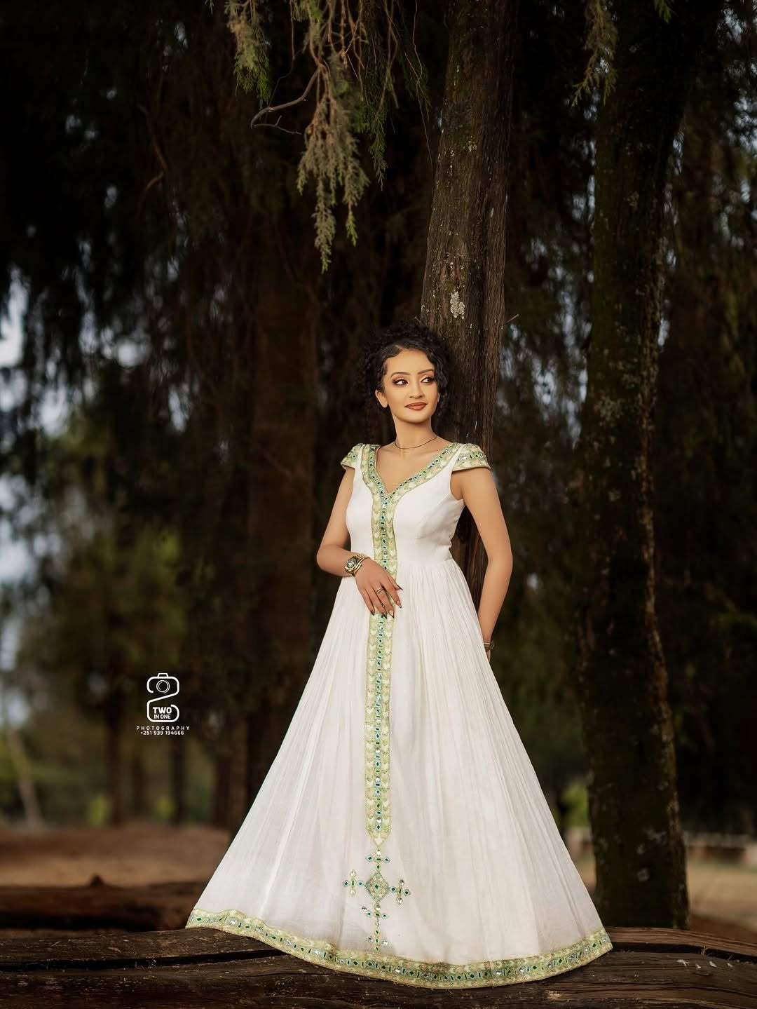 Woman in a white Green Ethiopian Traditional Dress with green embroidery standing outdoors against a tree.