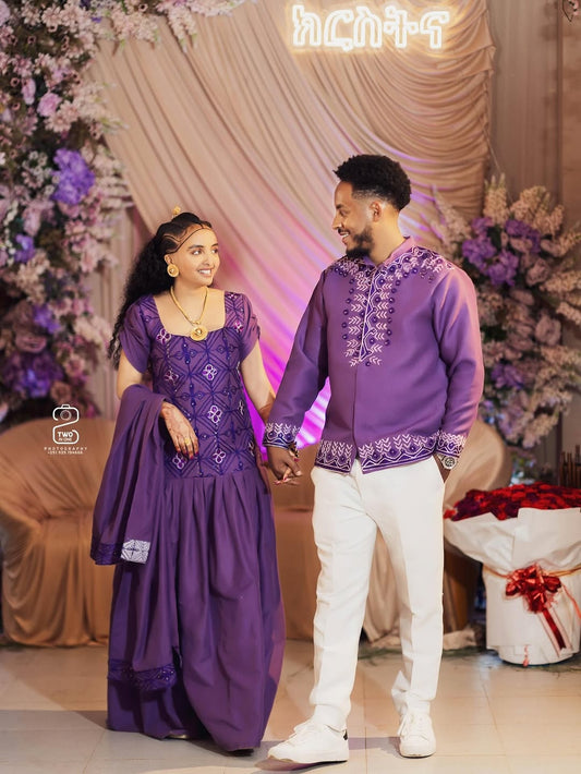 Man and woman in matching purple outfits standing in front of a decorated backdrop with flowers and gifts.