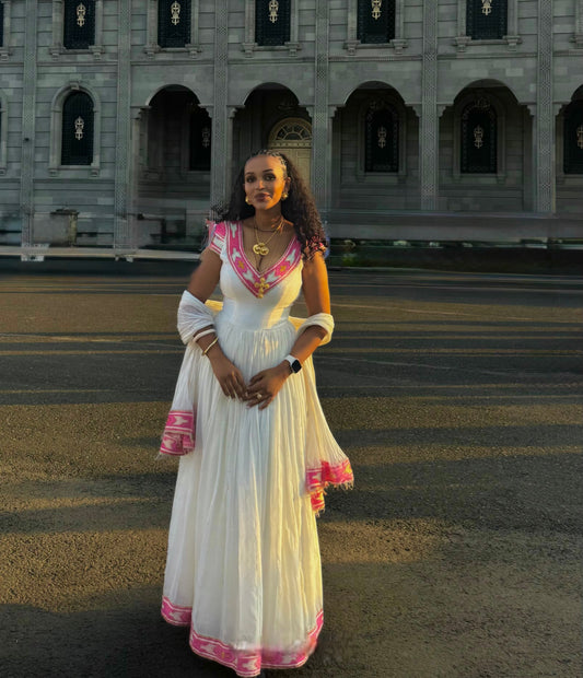A woman stands outdoors in front of a grand stone building, wearing a long white Ethiopian National Dresses with pink and gold embroidered details and draped sleeves.