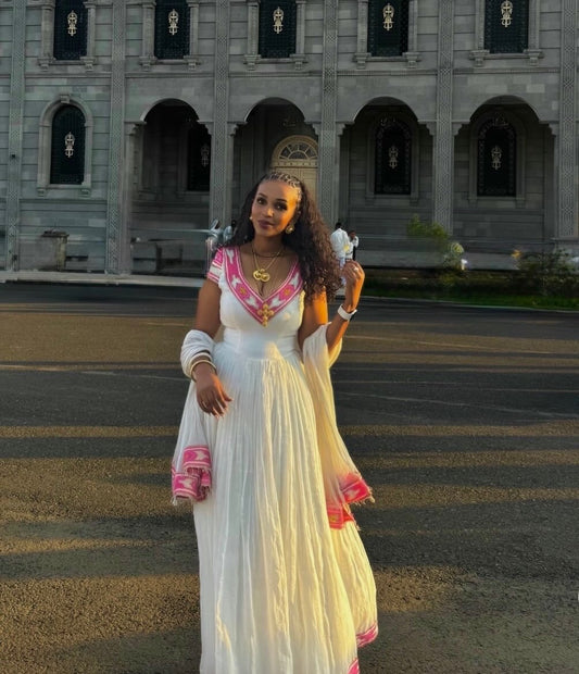 A woman wearing a white traditional Ethiopian National Dresses with pink and red embroidered details stands outdoors in front of a large gray stone building at sunset.