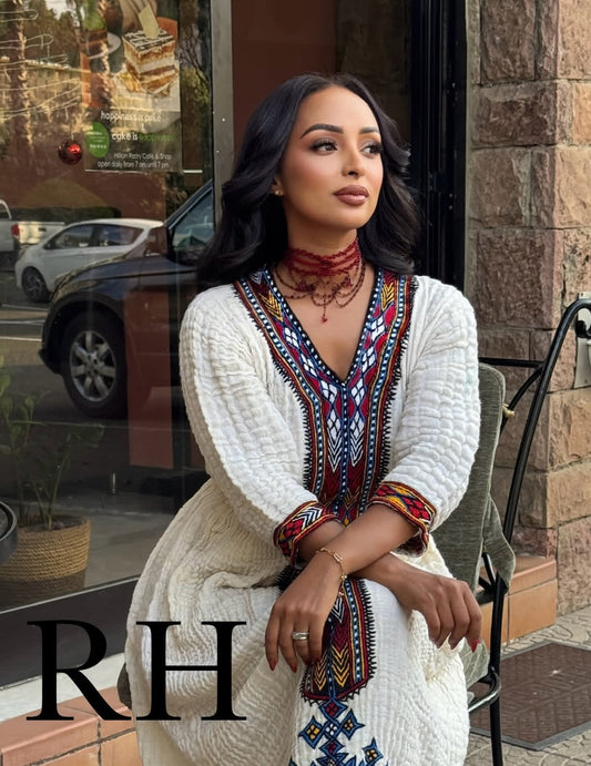 Woman in a white and patterned Traditional Ethiopian Dress sitting on a chair outside a building with 'RH' branding.