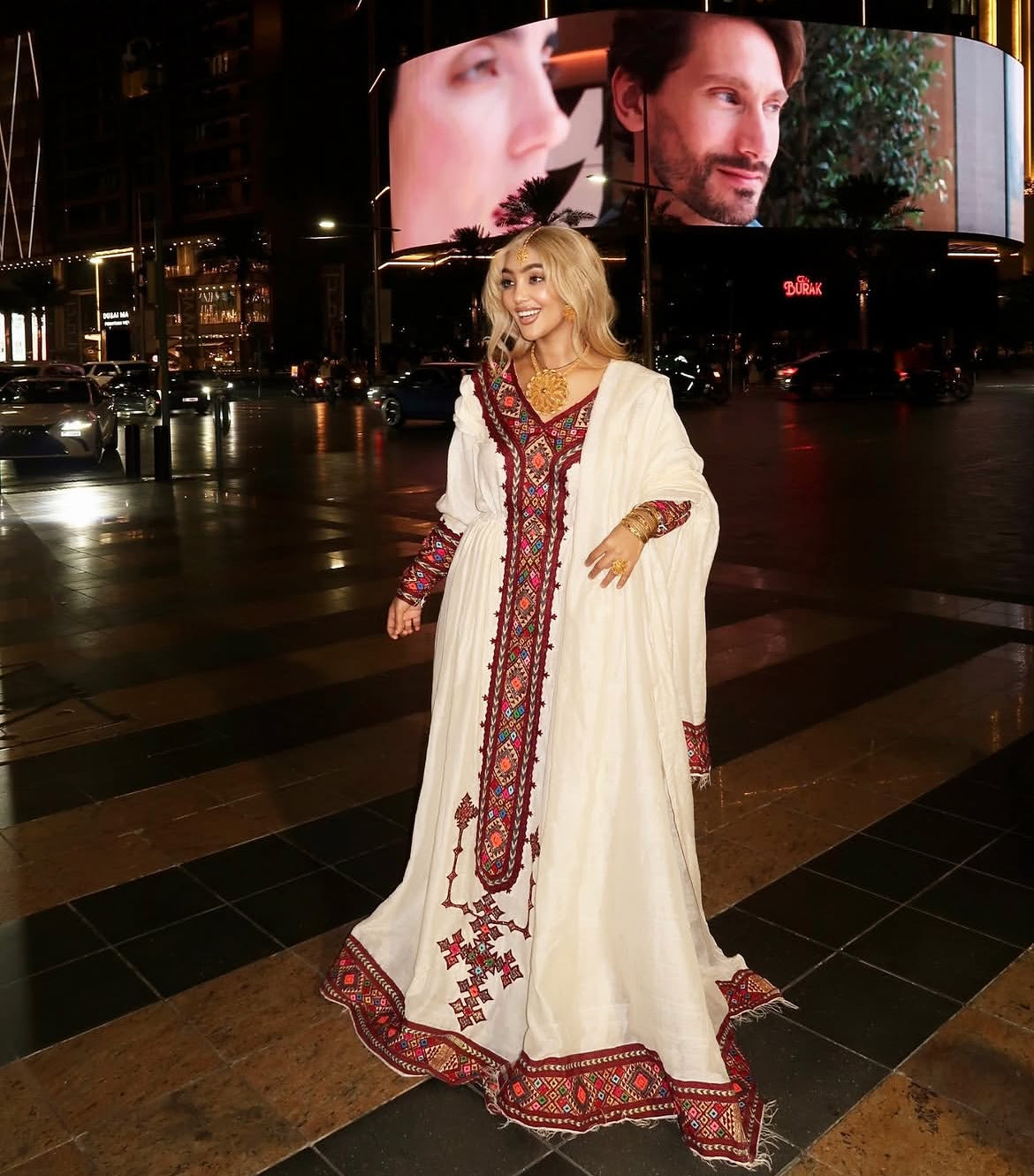 Woman in a white Habesha Dress with red and blue embroidery walking on a city street at night.