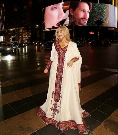 Woman in a white Habesha Dress with red and blue embroidery walking on a city street at night.