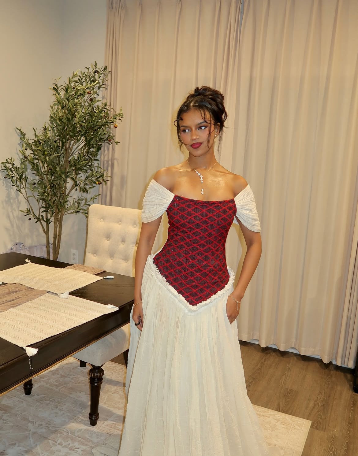Woman in a red and white habesha dress standing in an indoor setting with a table and plant.
