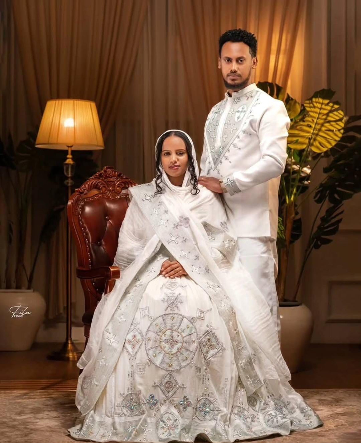 Man and woman in formal white attire posing together in a decorated room.