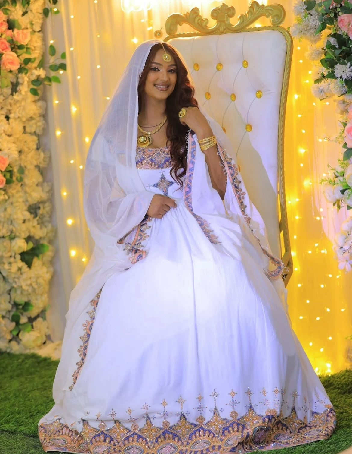 Woman in a white and gold traditional Habesha Dress sitting on an ornate chair with floral decorations and string lights in the background.