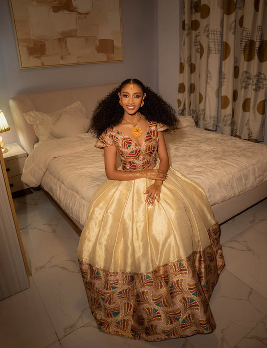 Woman in a patterned Habesha wedding Dress sitting on a bed in a bedroom.
