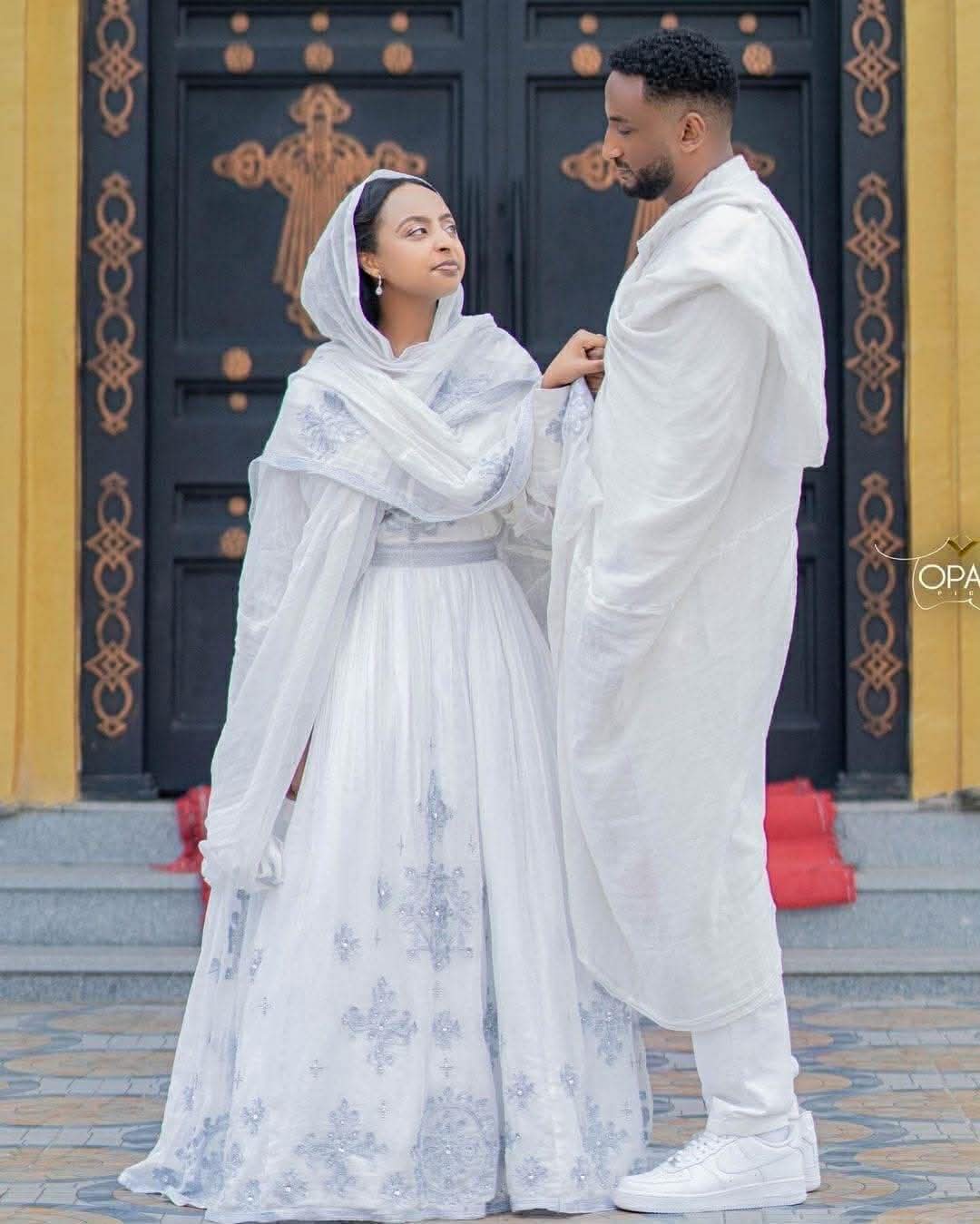 Couple in traditional white attire standing in front of a decorative door.