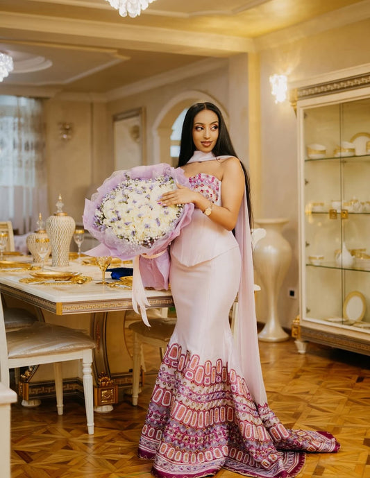 Woman in a pink and purple Habesha Dress holding flowers in an elegant room.