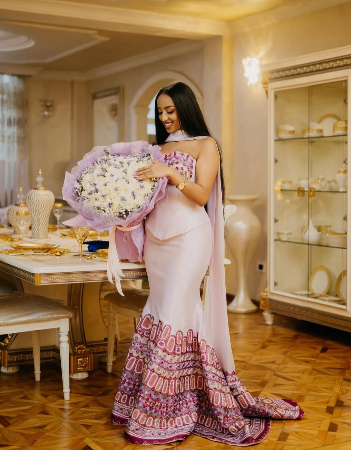 Woman in a elegant Habesha Dress holding a bouquet of flowers in a decorated room.