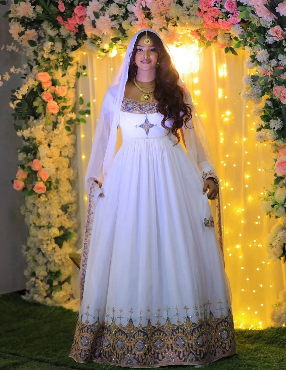 Woman in a white traditional Habesha Dress with gold details standing in front of floral decorations and string lights.