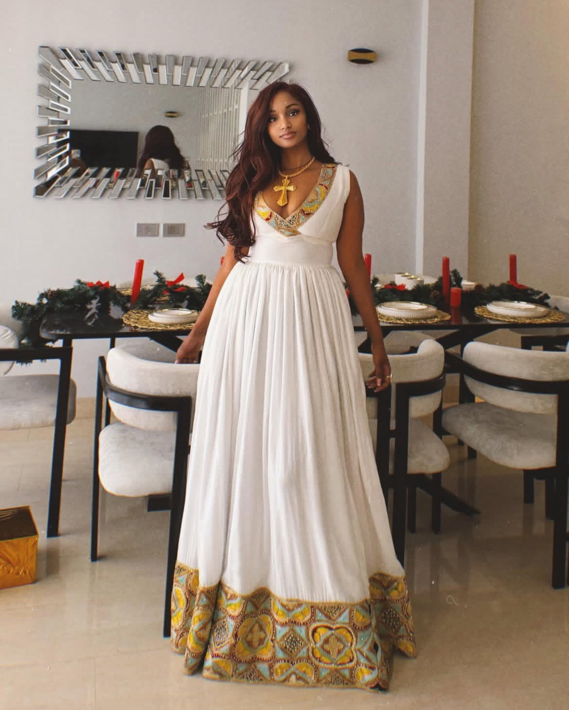 Woman in a white Habesha Zuria with gold patterns standing in a dining room.