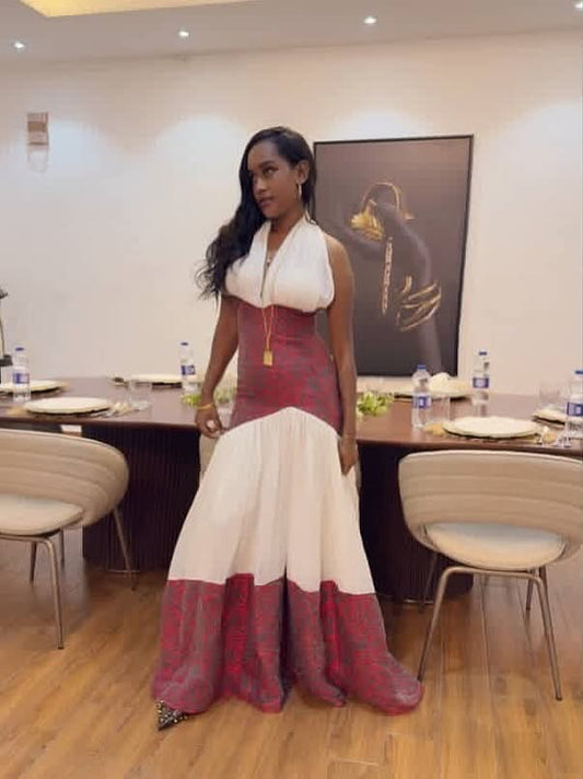 A woman wearing a formal floor-length Traditional Ethiopian Clothing with a white halter-style bodice and a voluminous skirt in white and deep red, standing in a dining or event room with tables and chairs.