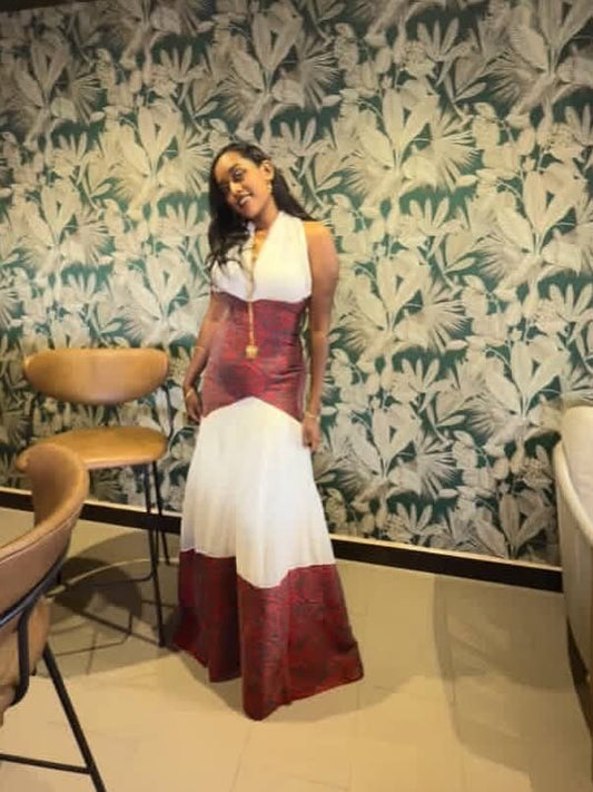 A woman wearing a sleeveless, color-blocked maxi Traditional Ethiopian Clothing with a white halter-style top, a deep red corset-style midsection, and a white and deep red layered skirt, standing in front of a green and white tropical leaf patterned wallpaper.