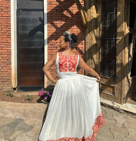 A woman stands outdoors in front of a brick and wooden building, wearing a sleeveless white Traditional Ethiopian Clothing with red geometric embroidery on the back and a matching red patterned hemline.