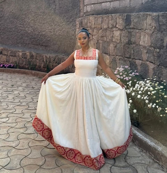 A woman wearing a traditional  white Traditional Ethiopian Clothing with coral-pink embroidered details and a matching embroidered hem, posing outdoors against a stone wall.