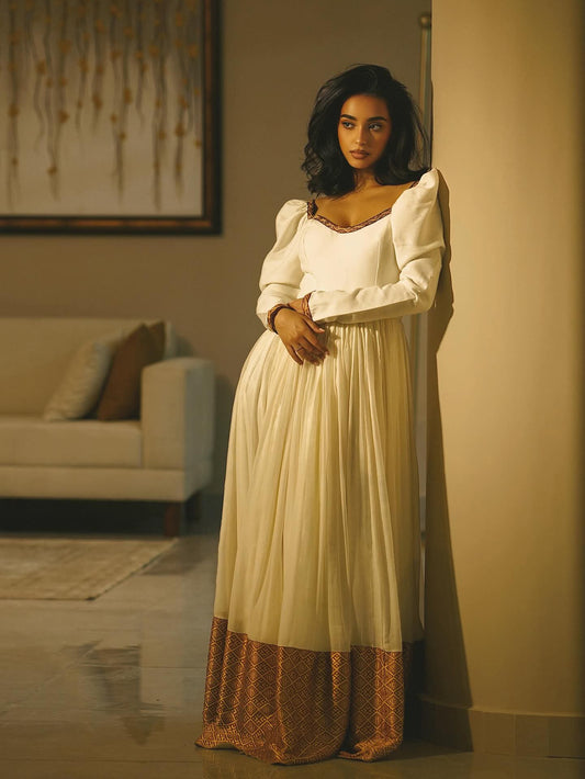 A woman wearing a long cream maxi Ethiopian Dress with puffed sleeves and a bronze patterned hem, standing in a warmly lit room next to a beige wall.