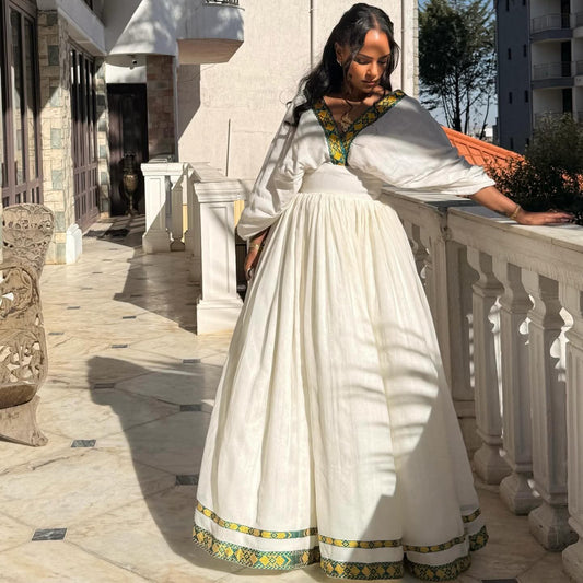 A woman wearing a traditional white long-sleeve Ethiopian Traditional Dress with green and yellow embroidered trim, standing on a sunlit stone balcony.