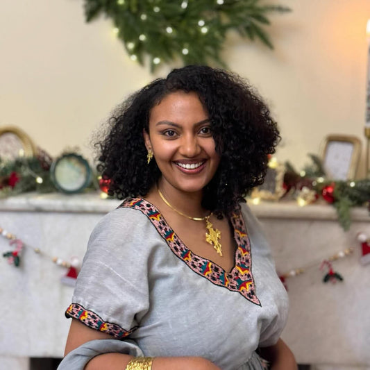 A smiling woman with curly black hair wearing a light Habesha Kemis with colorful embroidered trim, posed in front of a decorated Christmas fireplace.
