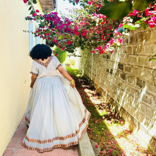 A woman wearing a traditional white Habesha Kemis with colorful embroidered details walks along a paved path next to a stone wall under a canopy of bright pink bougainvillea flowers.