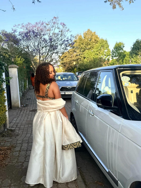Woman in a white Ethiopian Dress standing next to a car with trees and a clear sky in the background