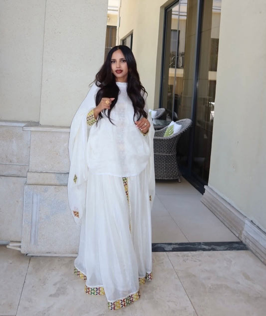 A woman wearing a white traditional Habesha Zuria with gold embroidered details, standing on a tiled patio in front of a beige building wall.