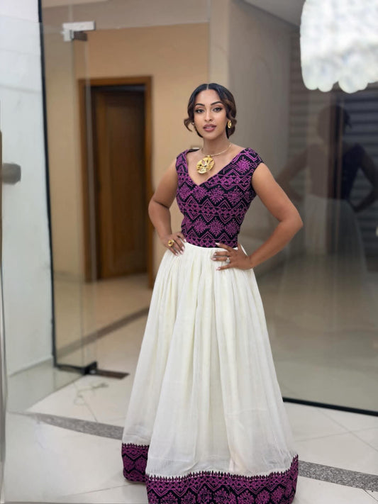 A woman modeling a sleeveless white maxi Ethiopian Dress with a purple embroidered bodice and a matching purple border on the skirt, standing in an indoor hallway with glass doors.