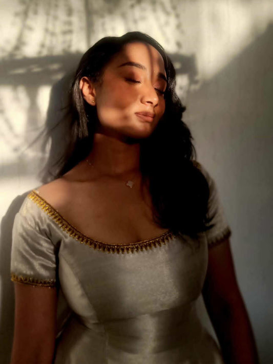 A woman wearing a short-sleeved satin Ethiopian Traditional Dress  with gold trim along the neckline and sleeve cuffs, standing against a wall with dramatic shadow light.