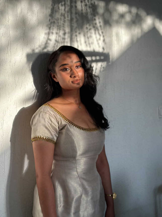 A woman with long dark hair stands against a textured white wall, wearing a light beige or off-white Ethiopian Traditional Dress  with gold trim along the neckline and sleeve cuffs. Strong natural shadows from a hanging plant are cast across the wall and her face.