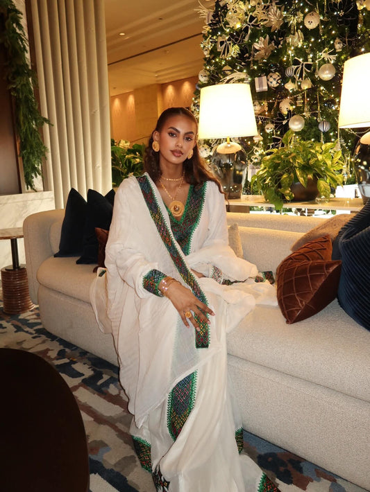Woman in a white Ethiopian Dress with green patterns sitting on a couch in a decorated room.