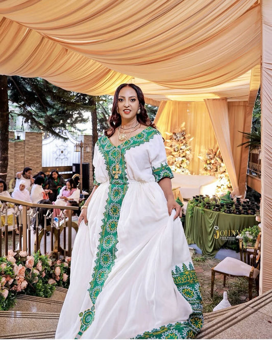 A woman wearing a white traditional Habesha Dress with green and gold embroidered details walks down outdoor steps under a draped beige canopy. Guests and tables are visible in the background at what appears to be an event or ceremony.