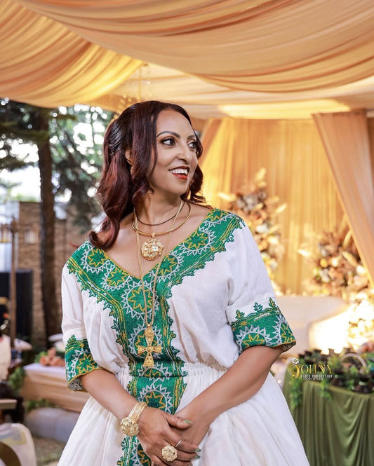 A woman wearing a white Habesha Dress with green and gold embroidered patterns, standing under a draped beige tent at an outdoor event.