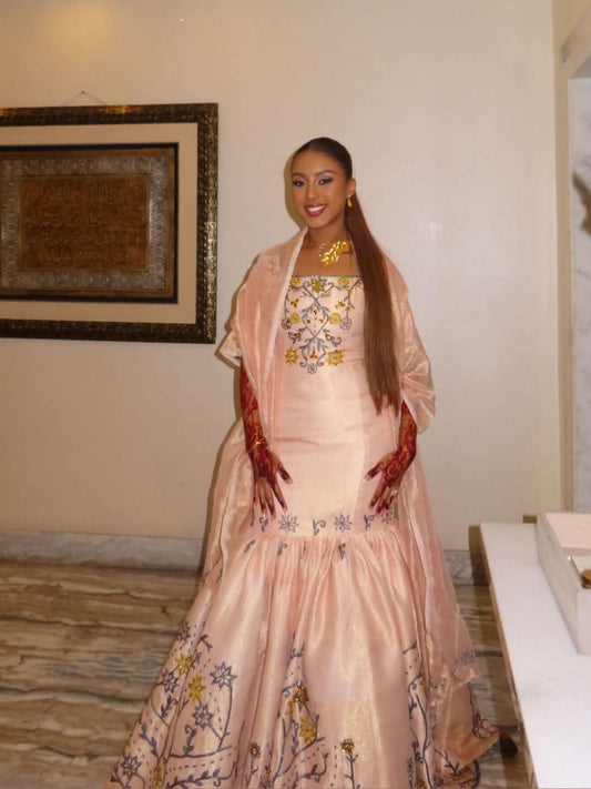 A woman wearing a traditional light pink Ethiopian Dress with a matching sheer shawl, standing in an indoor setting with a beige wall and framed artwork in the background.