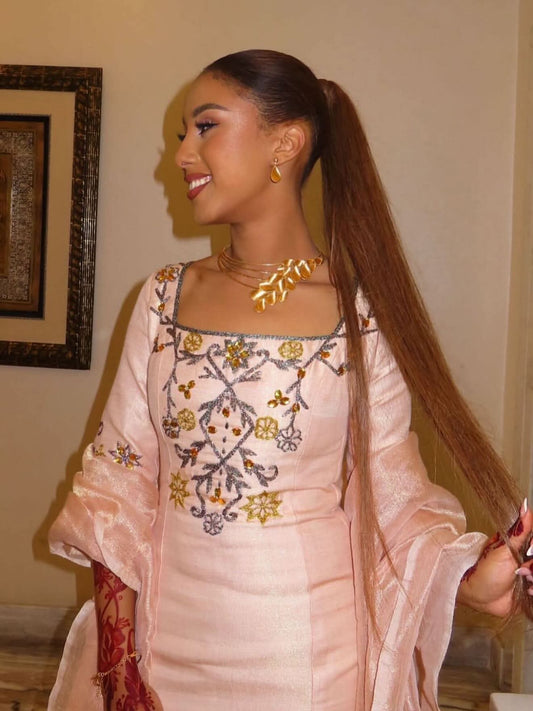 A woman wearing a light pink embroidered traditional Ethiopian Dress with gold and gray detailing, a matching pink dupatta, and gold jewelry, standing indoors against a beige wall.
