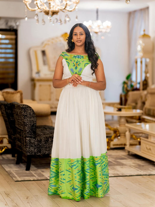 A woman modeling a floor-length white Traditional Ethiopian Clothing with vibrant green and blue patterned embroidery on the bodice and hem, standing in a furnished indoor room with chandeliers and furniture.