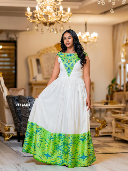 A woman modeling a sleeveless white maxi Traditional Ethiopian Clothing with a green and blue geometric patterned yoke and hem, standing in an ornate, luxurious room with gold chandeliers.