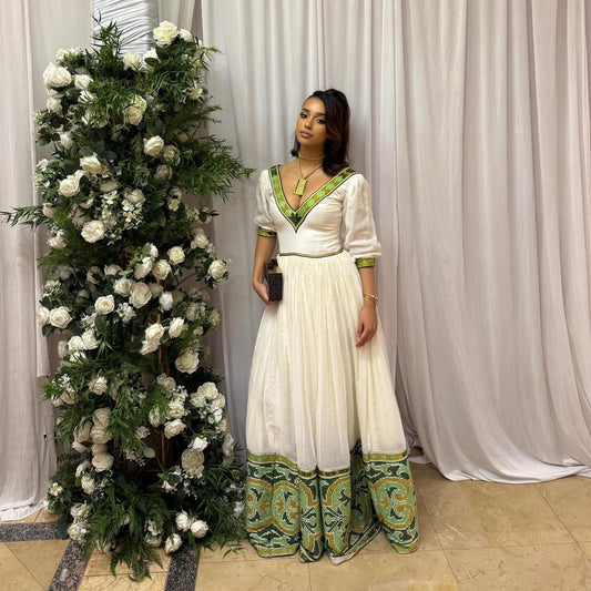 A woman wearing a white Habesha Dress with green embroidered details stands next to a large floral arrangement in front of white draped curtains.
