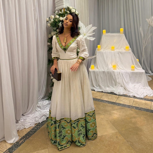Woman in a white and green Habesha Dress standing in front of a decorated setup with candles and flowers.