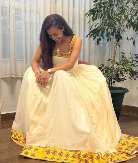 A woman wearing a cream-colored traditional Habesha Zuria with yellow and red embroidered borders, sitting on a chair in an indoor setting with curtains and a potted plant in the background.