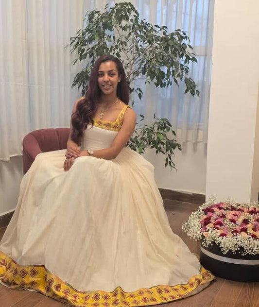 A woman wearing a traditional Indian white or cream Habesha Zuria with gold embroidery, sitting on a pink armchair in an indoor setting with a potted plant and a flower arrangement in the background.